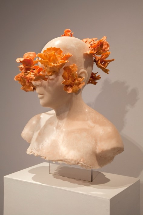 A stone carved bust of a bald figure. Out of front, side, and back of head are leaf-like structures