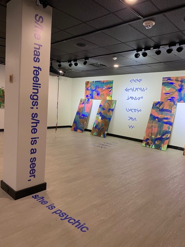 Interior of gallery, paintings hang on and lean against wall. English and Indigenous text on walls and floor