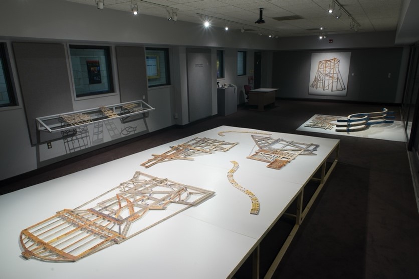 Gallery room with white tables displaying wooden architectural models under soft lighting, with additional pieces mounted on the walls
