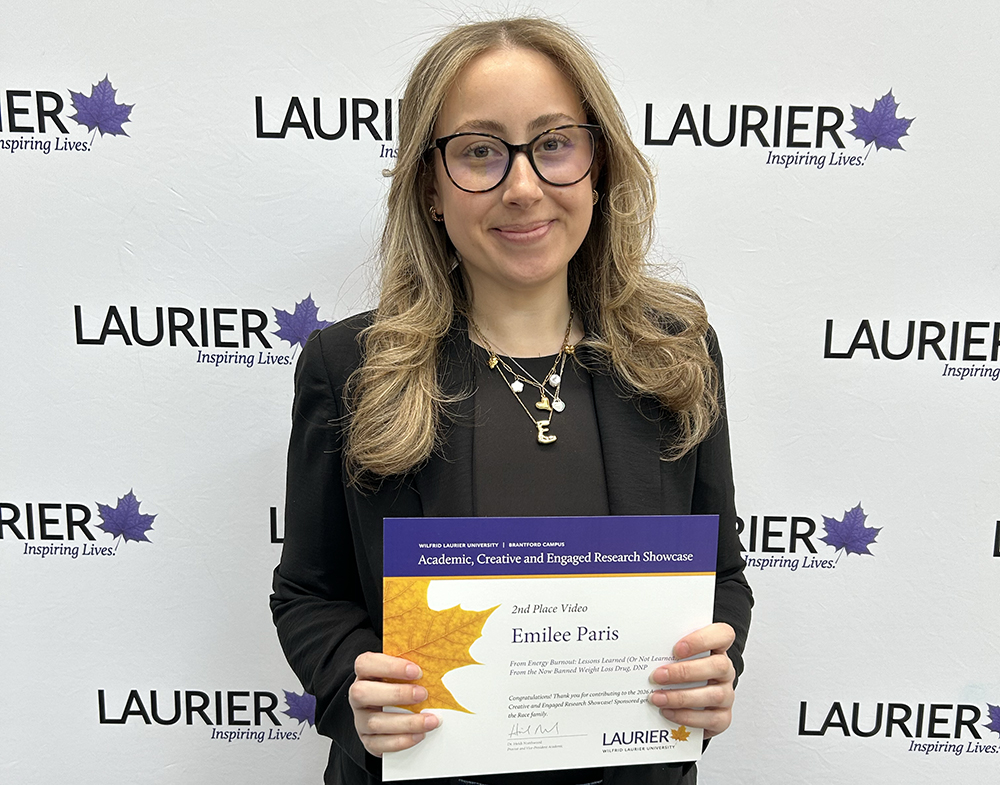 Emilee paris holds her ACERS certificate in front of a Laurier-branded backdrop