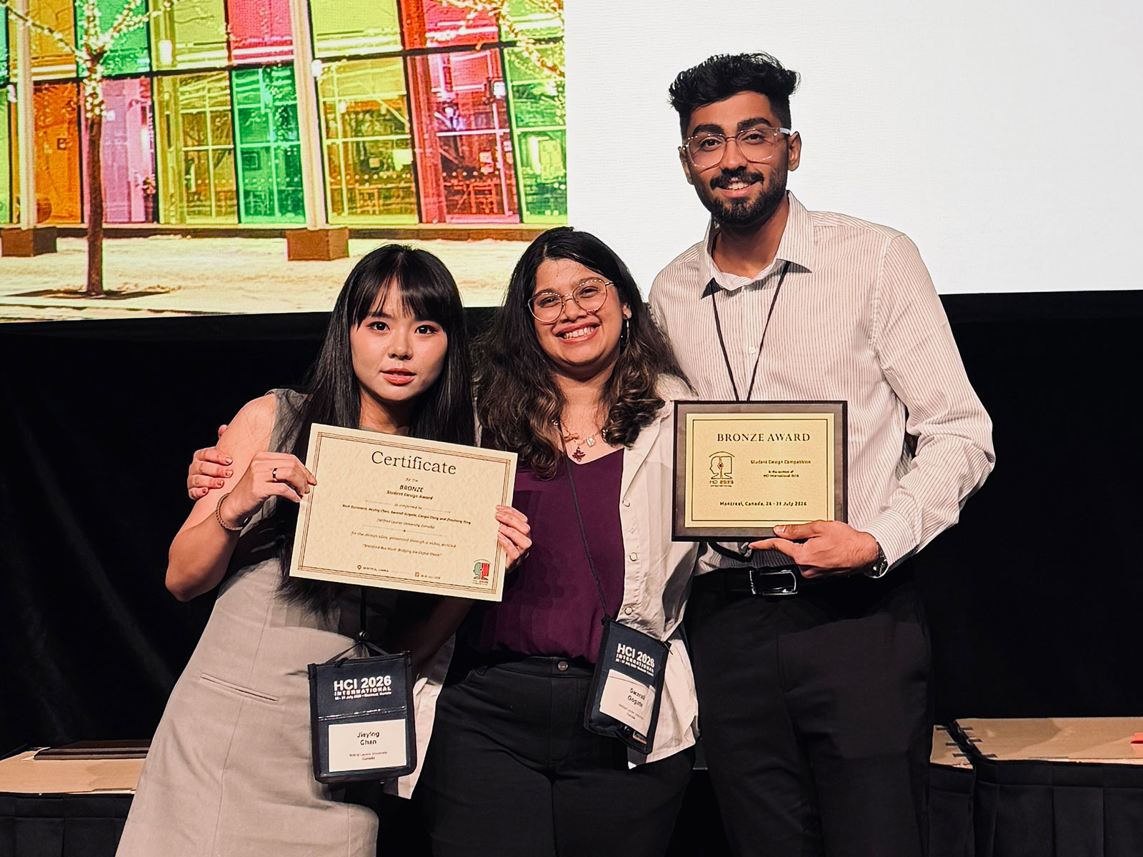 Three students stand on a stage holding bronze award plaques and certificates