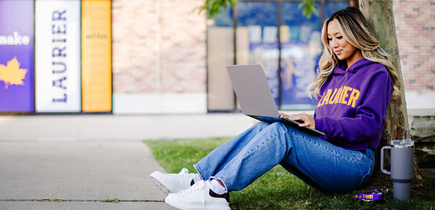 female student in purple laurier sitting on grass smiling at laptop
